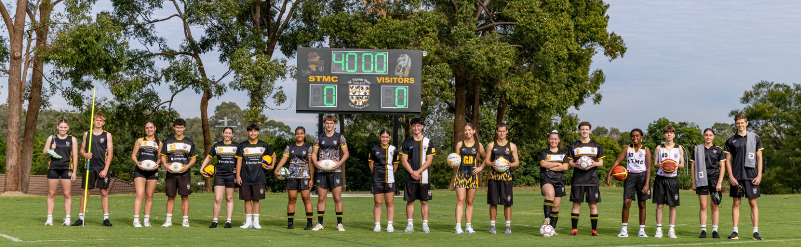 students standing together on a sports field holding a variety of sporting equipment in front of a school scoreboard, highlighting diverse sporting opportunities, participation and an inclusive school sports culture