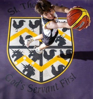 student jumping with a basketball above the college crest on a court, highlighting energy, skill and engagement in sport