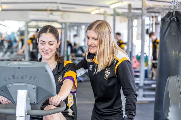 student using a cardio machine in a fitness centre with staff support, highlighting wellbeing, training and guided physical development